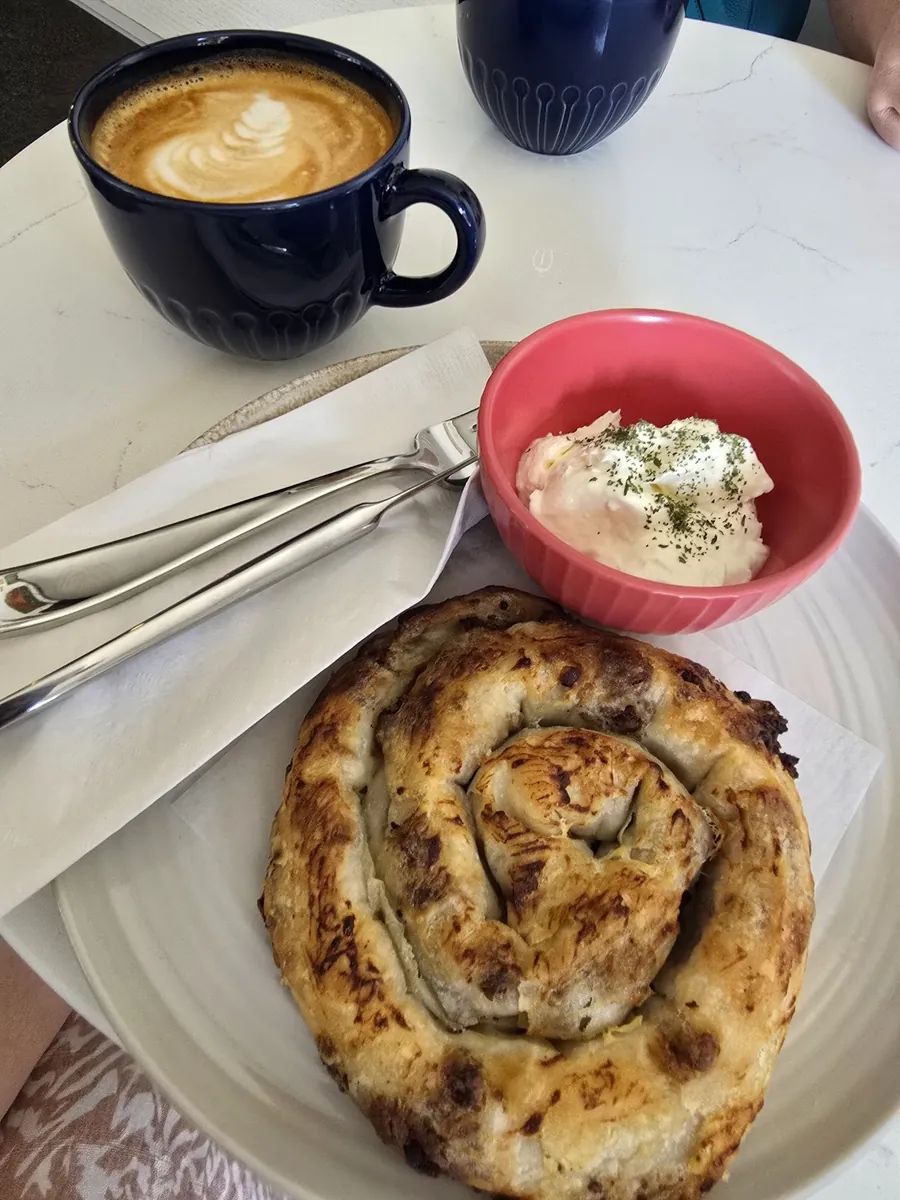 A golden spiral of börek on a white plate, beside a pink bowl of white cheese and a blue mug of coffee
