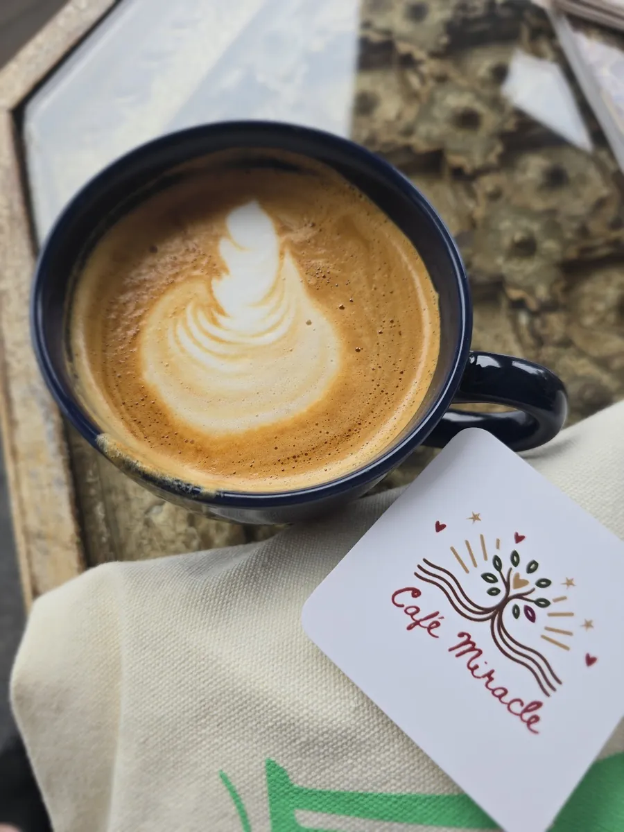 A latte with leaf-pattern foam in a deep blue ceramic mug, on a tote bag printed with the Café Miracle logo