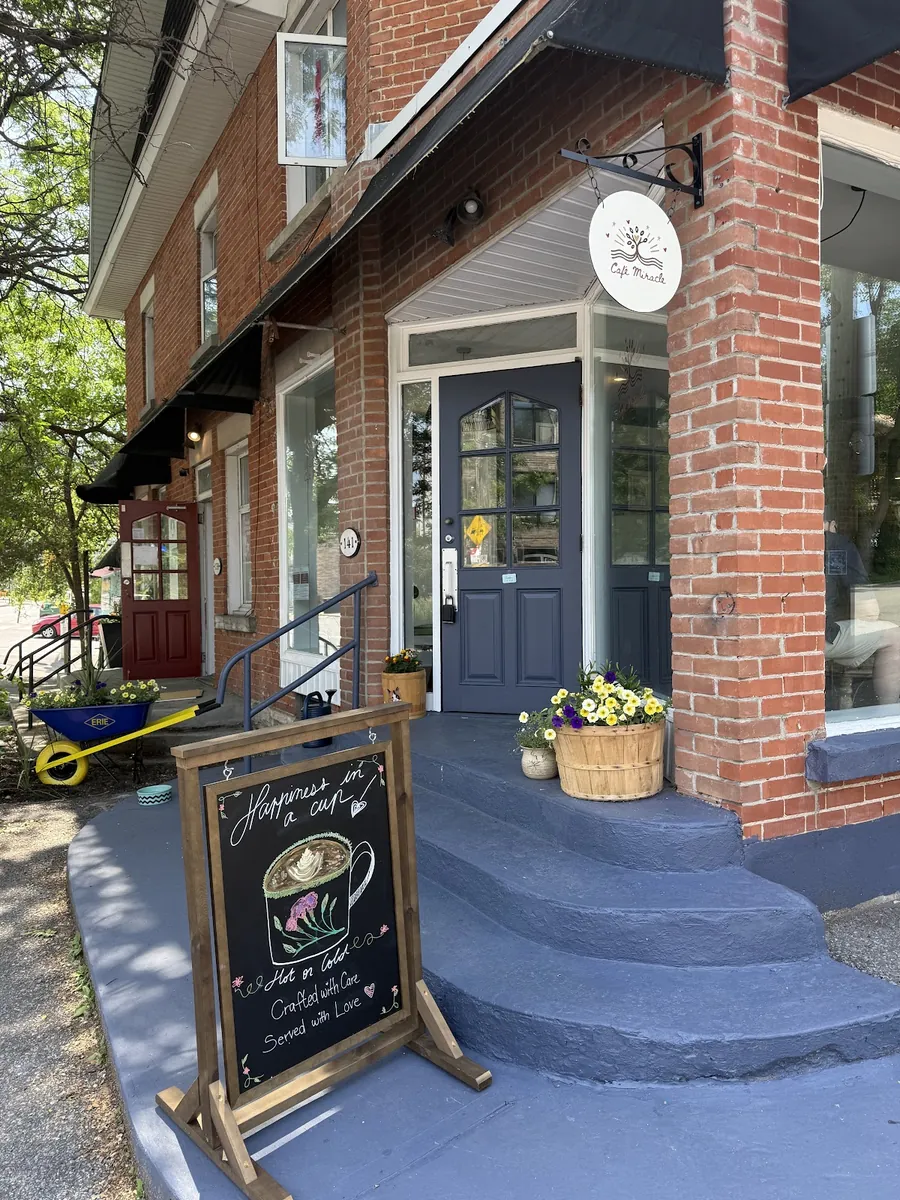 The brick front of Café Miracle on Beechwood Avenue, with a blue door, blue steps, flower pots and a chalkboard sign